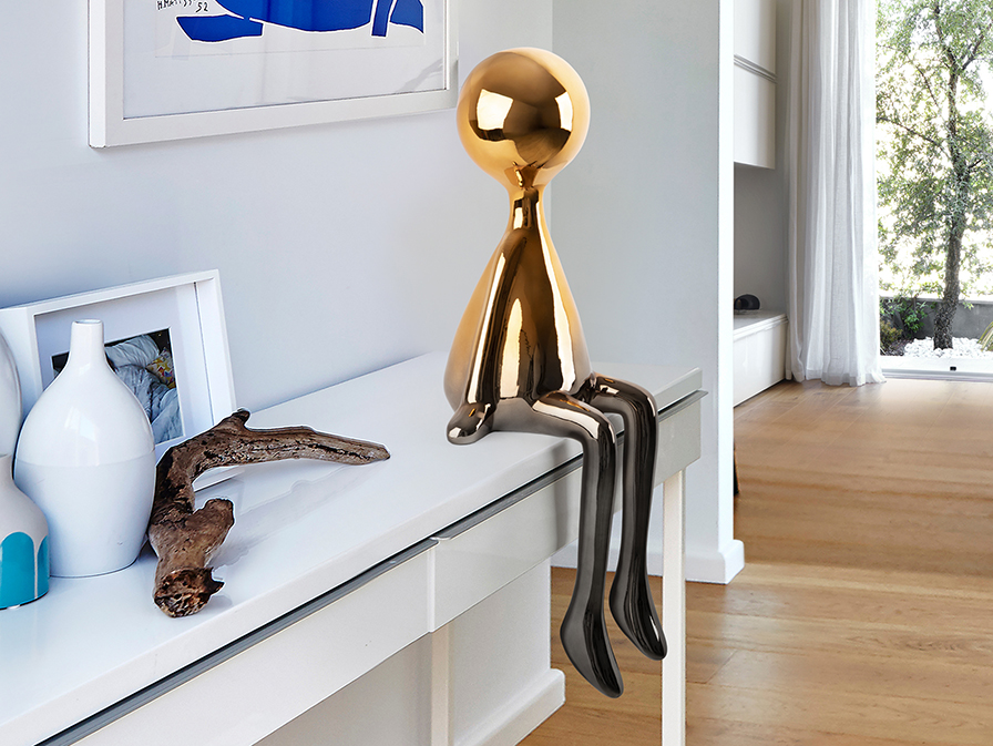 Metallic seated sculpture with a rounded orange-toned head and long grey legs hanging over a console